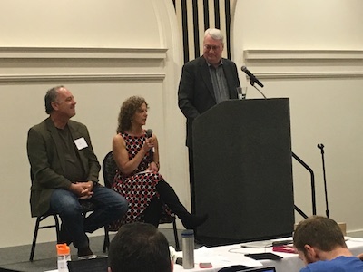 Conference image with two seated panelists and a moderator standing nearby