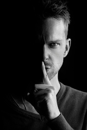 Man in shadows holding up a finger to his lips to indicate silence or secrets